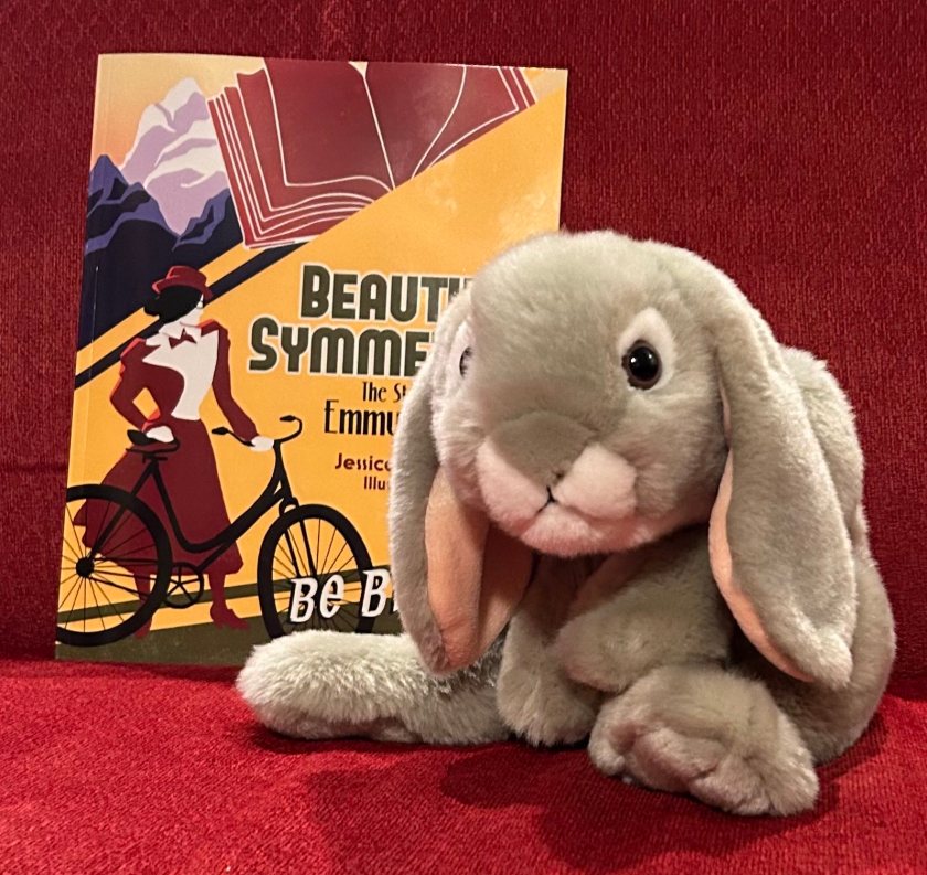 Sprinkles is posing with Beautiful Symmetry: The Story of Emmy Noether, written by Jessica Christianson and illustrated by Brittany Goris.
